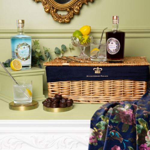 Brown wicker hamper surrounded by a bottle of sloe gin and dry gin. There is a jar of lemons and limes on top of the hamper, next to a glass of gin and tonic with a metal straw. At the front of the hamper is a tray of rose and violet creams, next to a sec