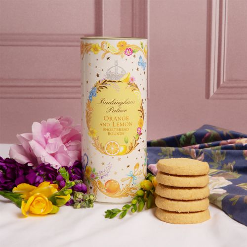 A cardboard tube of orange and lemon biscuits. There is a floral garland at the top and bottom of the tube. At the centre of the tube is an orange oval reading the words 'Buckingham Palace' in gold and 'Orange and lemon shortbread rounds' in orange. 