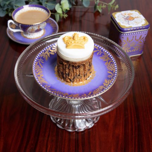 Round fruit cake topped with white icing and gold icing crown. The cake is stood in front of a cube tin designed with the coat of arms and purple and gold Platinum Jubilee design of gold leaf and crown