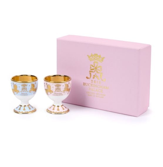 Two egg cups, both with a lion and unicorn design, one blue with "Prince George" and one pink with "Princess Charlotte" in gold. The egg cups are positioned next to a pink presentation box. Two egg cups, both with a lion and unicorn design, one blue with "Prince George" and one pink with "Princess Charlotte" in gold. The egg cups are positioned next to a pink presentation box.