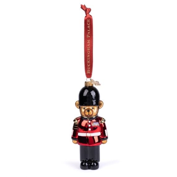 Glass Christmas ornament shaped like a King's Guard in traditional uniform, featuring a bright red tunic, black trousers and a black bearskin-style hat. The ornament hangs from a red ribbon printed with “BUCKINGHAM PALACE” in gold lettering.