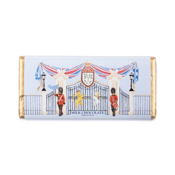Milk chocolate bar wrapped in a pale blue sleeve illustrated with decorative gates, royal guards, and heraldic motifs, finished with gold foil ends and the text “The Royal Sweet Shop” and “Milk Chocolate – Made in the UK”.
