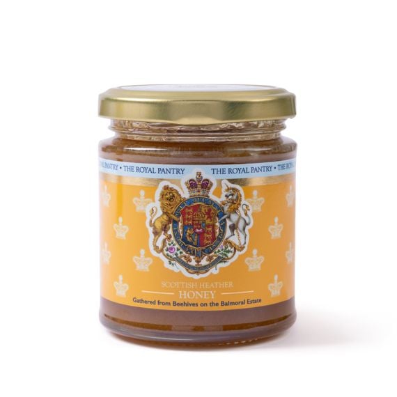 Front view of a glass jar of Scottish Heather Honey from The Royal Pantry, featuring a gold lid and an amber label decorated with the Royal Coat of Arms, including a lion and unicorn. The honey is gathered from beehives on the Balmoral Estate.