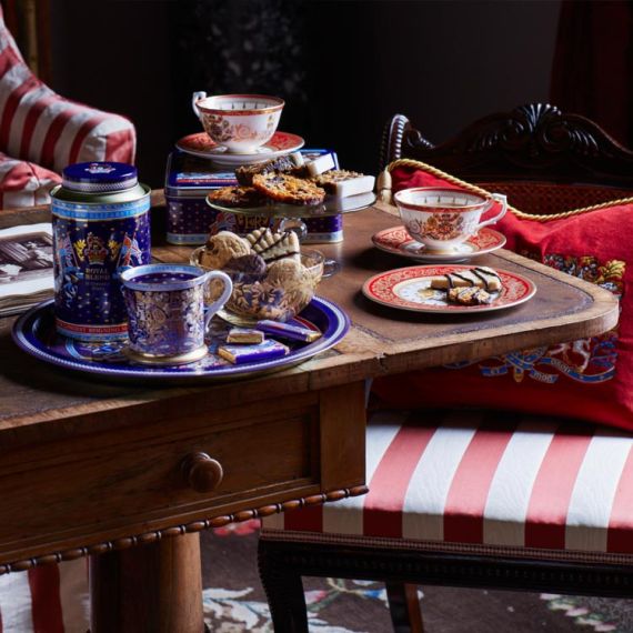 The longest reigning monarch tea set on a wood table. Biscuits are in a dish and on a plate. There is a red cushion on a chair 
