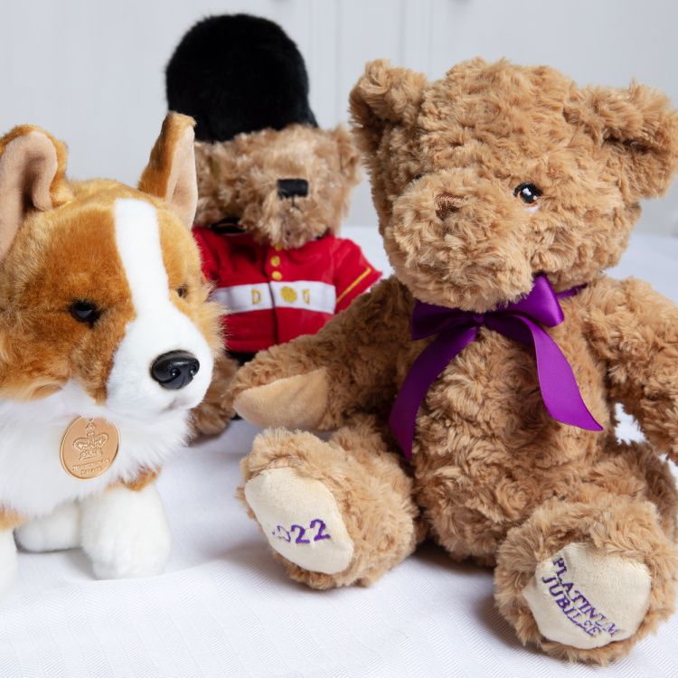 Brown teddy bear with a purple ribbon tied round its neck. Its feet are embroidered with 2022 in purple thread and on the other foot is 'Platinum Jubilee' embroidered in purple threads. Next to the bear is a bear dressed as a guardsman and a corgi with a 