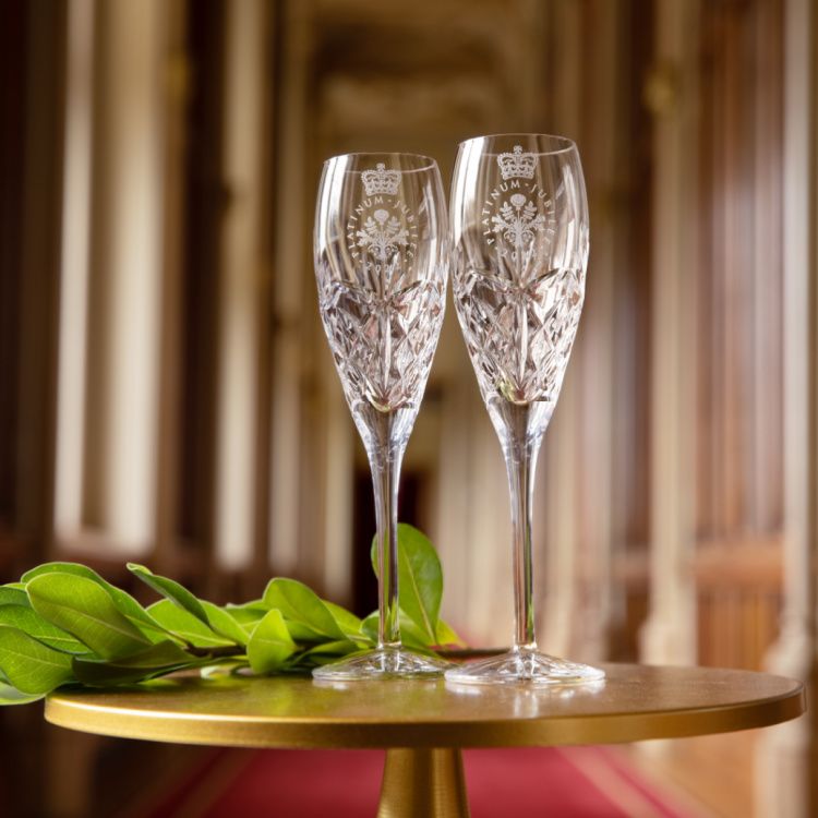 Two crystal champagne glasses etched with the Platinum Jubilee emblem on a gold table with greenery. The glasses are placed in front of a long corridor.