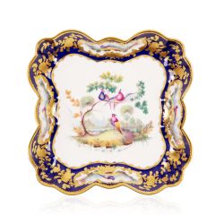 Square dish with a gold and blue ornate border. At the centre of the plate is a painted design of three exotic birds in a tree.