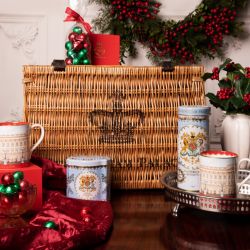 Creative of the wicker hamper surrounded by a blue tea caddy, blue biscuit tube. On a tray is a mug featuring Buckingham Palace. The same mug also features to the left of the hamper stood on a red box. There is a bowl of red and green foiled truffles to t