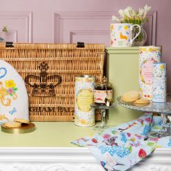 Wicker hamper on a pink panel background surrounded by eucalyptus, printed mug, gold spoon, floral box of tea and floral tube of biscuits and some biscuits scattered in front of the hamper