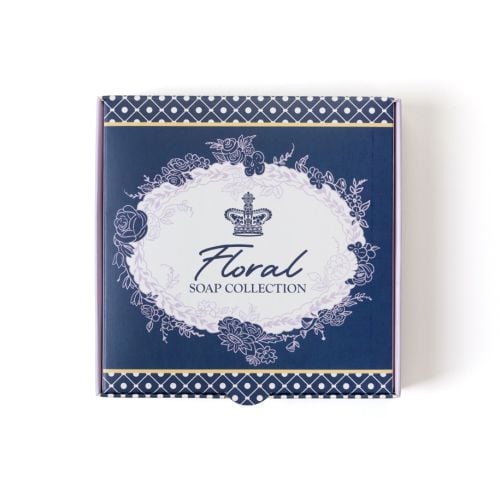 Open pastel purple gift box displaying a set of nine square soaps arranged in a 3x3 grid, each in different soft colours including pink, green, beige, yellow, and lavender. The inside lid features a decorative floral wreath design with descriptive text ab