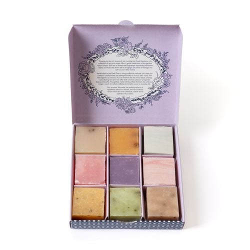 Open pastel purple gift box displaying a set of nine square soaps arranged in a 3x3 grid, each in different soft colours including pink, green, beige, yellow, and lavender. The inside lid features a decorative floral wreath design with descriptive text ab